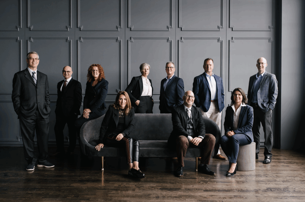 A professionally dressed group of eleven business professionals posing together in a modern studio. Some are standing while others are seated on a dark sofa and a small stool. They wear suits and business-casual attire, and the background features a gray paneled wall with a dark wood floor, creating a formal and polished team portrait atmosphere.