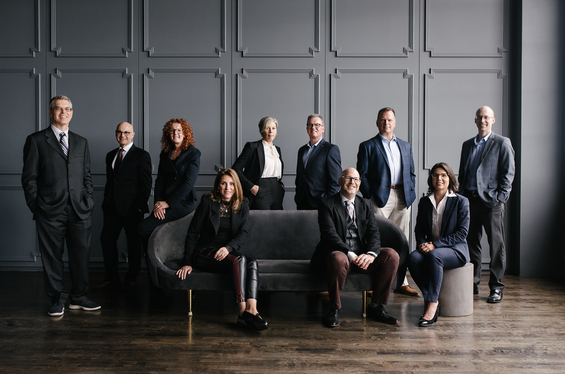 A professionally dressed group of eleven business professionals posing together in a modern studio. Some are standing while others are seated on a dark sofa and a small stool. They wear suits and business-casual attire, and the background features a gray paneled wall with a dark wood floor, creating a formal and polished team portrait atmosphere.