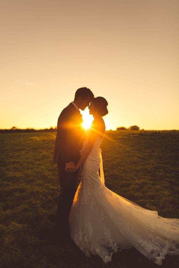A silhouetted couple standing close together in an open field at sunset, holding hands with their foreheads gently touching. The sun is low on the horizon behind them, creating a warm glow and highlighting a flowing wedding dress and a formal suit, giving the scene a romantic, serene atmosphere.