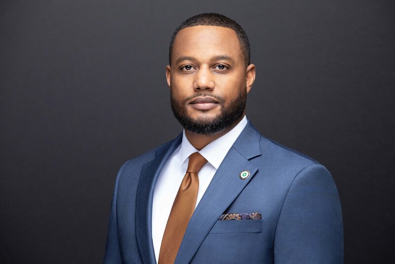 A professional head-and-shoulders portrait of a man wearing a blue suit, white dress shirt, and brown tie. He has a neatly trimmed beard and short hair, and is looking directly at the camera with a calm, confident expression. The background is dark and neutral, emphasizing a polished, formal appearance.